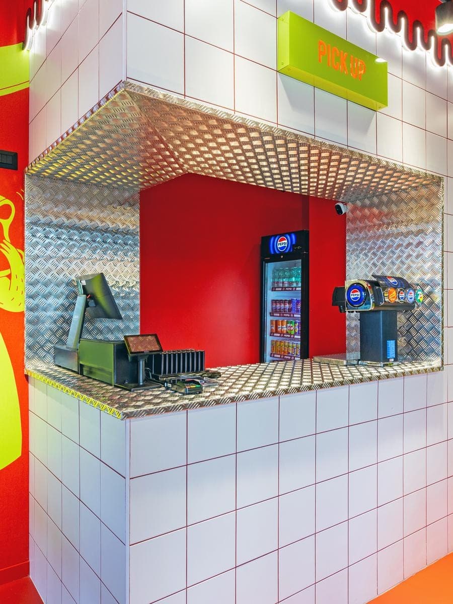Banco pick-up di un punto vendita Cucchi Burgers con pareti rosse, piastrelle bianche e mascotte lime del macellaio
