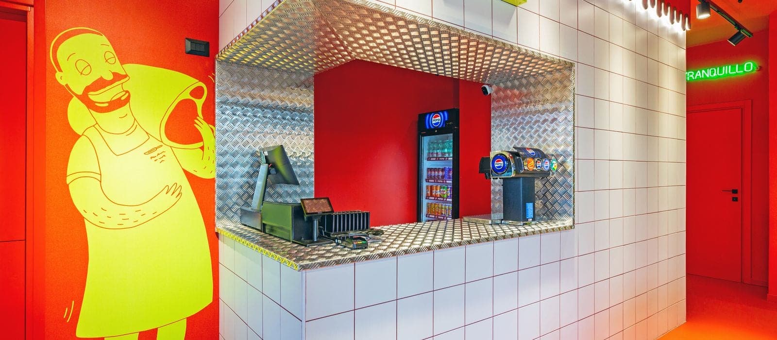 Banco pick-up di un punto vendita Cucchi Burgers con pareti rosse, piastrelle bianche e mascotte lime del macellaio