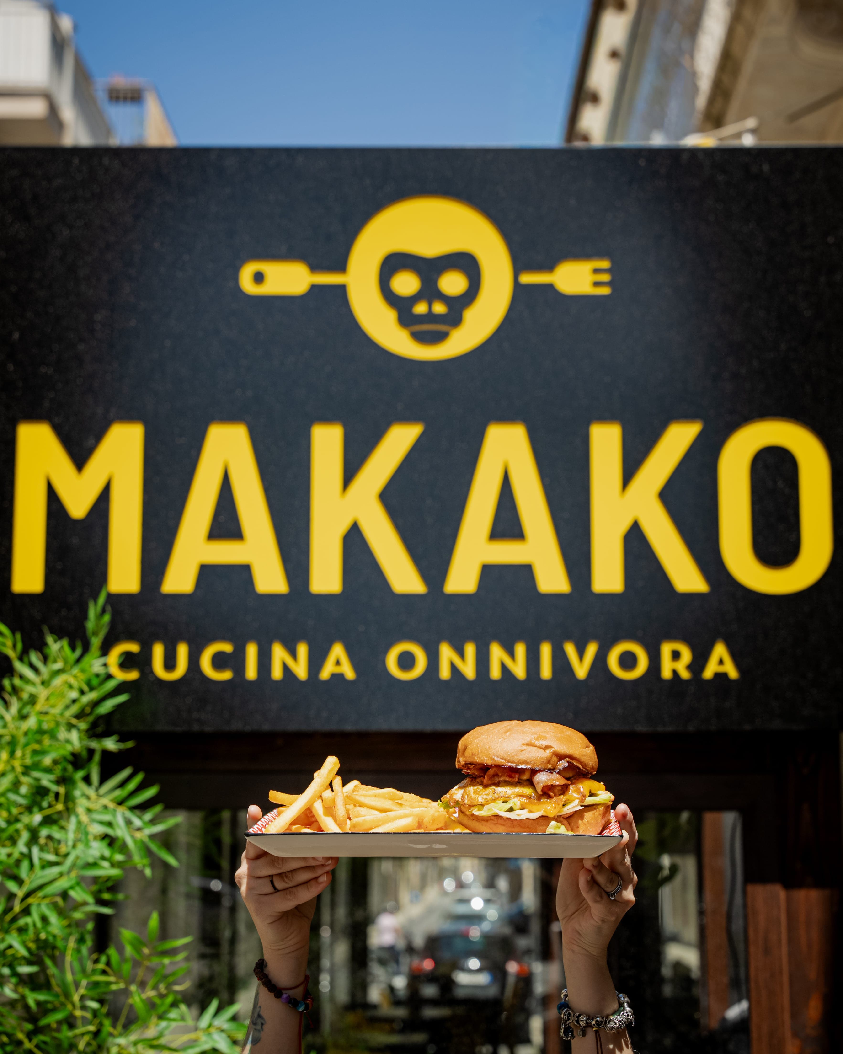 Ristoratore Makako Cucina Onnivora con il grembiule nero del brand e braccia incrociate, davanti a un blocco giallo