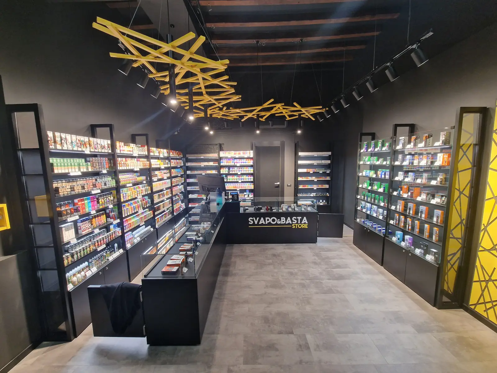 Interno di uno Svapo&Basta Store: pareti di prodotti, banco con l'insegna e soffitto a travi gialle